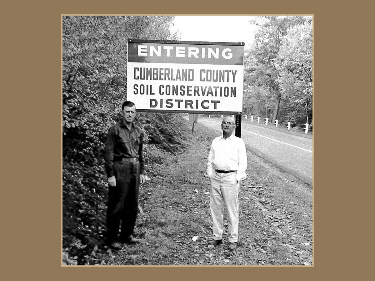 Black and white photo of 2 men standing by Conservation District sign along road